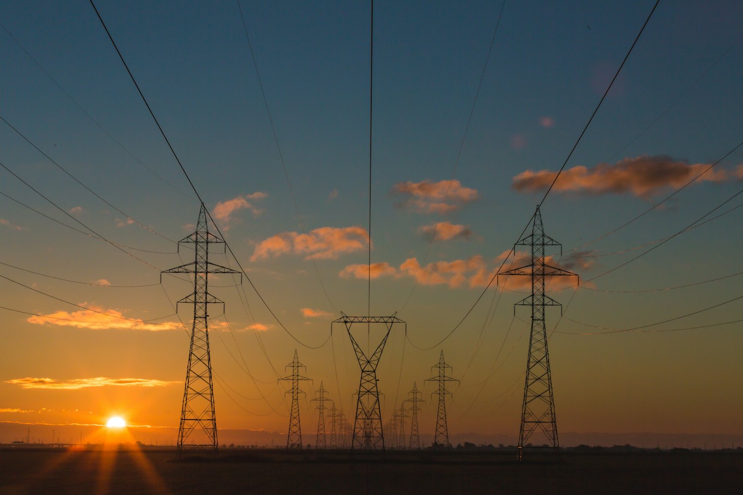 Silhouetted high-voltage transmission towers and power lines against an orange sunset